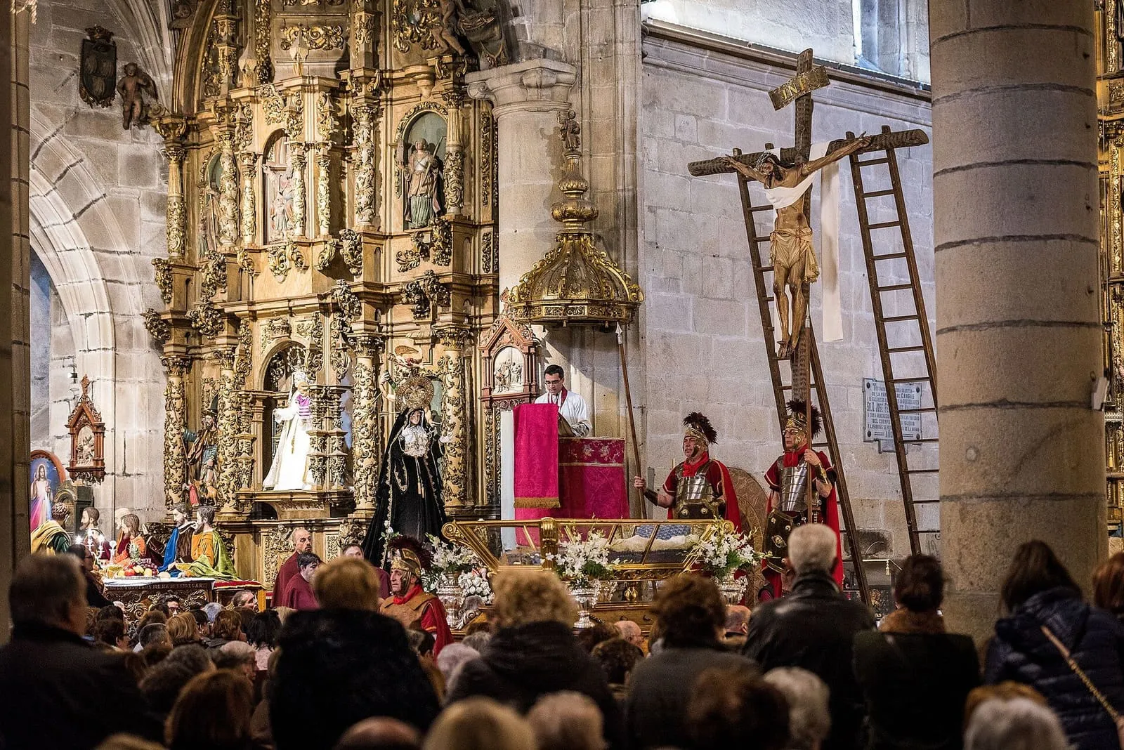Una ceremonia de Semana Santa en la excolegiata de Cangas, con cofrades de rojo, centuriones romanos y el Cristo en la cruz con escaleras, ante el retablo dorado