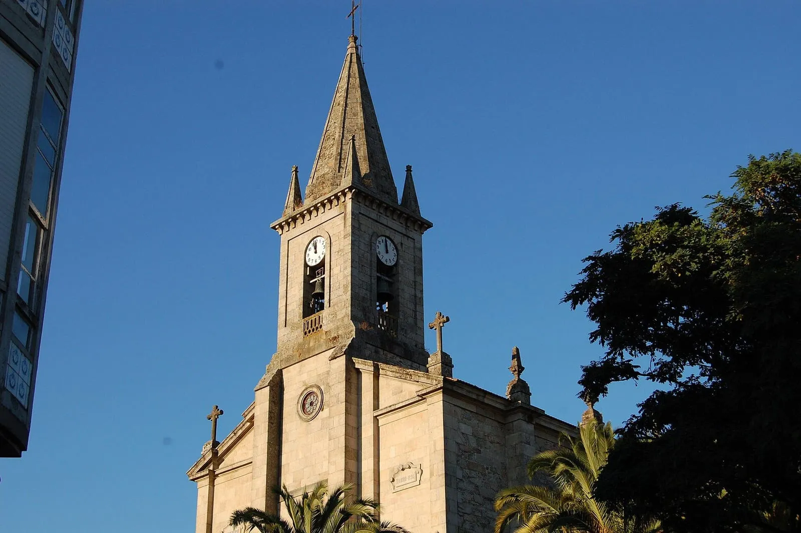 La iglesia de Santo Tomé de Caldas, de piedra clara, con su torre con aguja y palmeras en primer plano