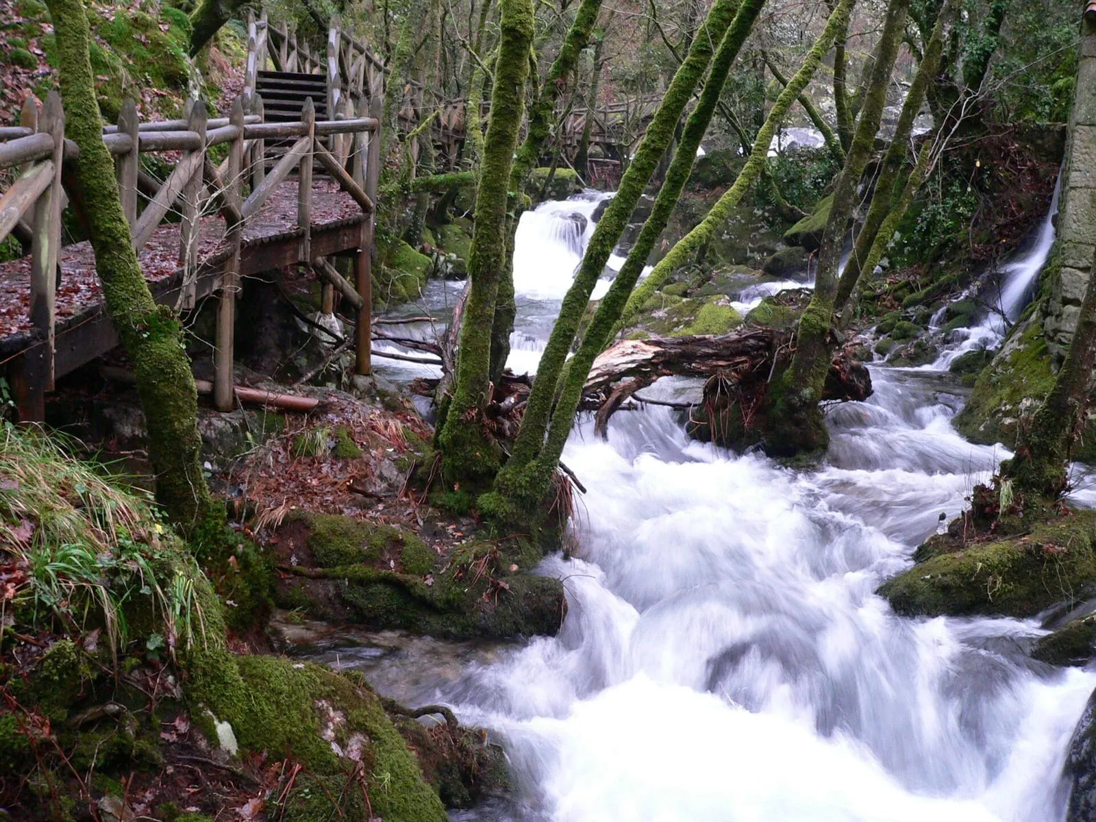 Cascada entre rocas y musgo con una pasarela de madera junto al río, en Valga