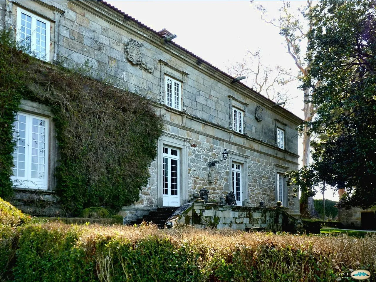 Fachada principal de granito del pazo de Rubiáns, con ventanas blancas y hiedra, tras un seto