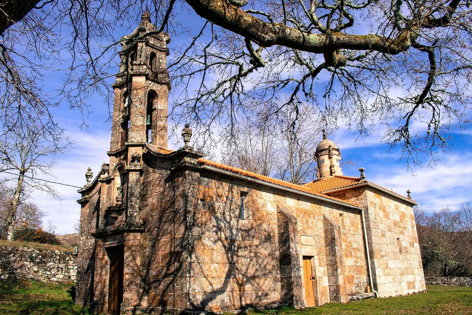 La capela da Virxe de Gundián, de piedra, con campanario y cubierta de teja, entre robles sin hojas y cielo azul