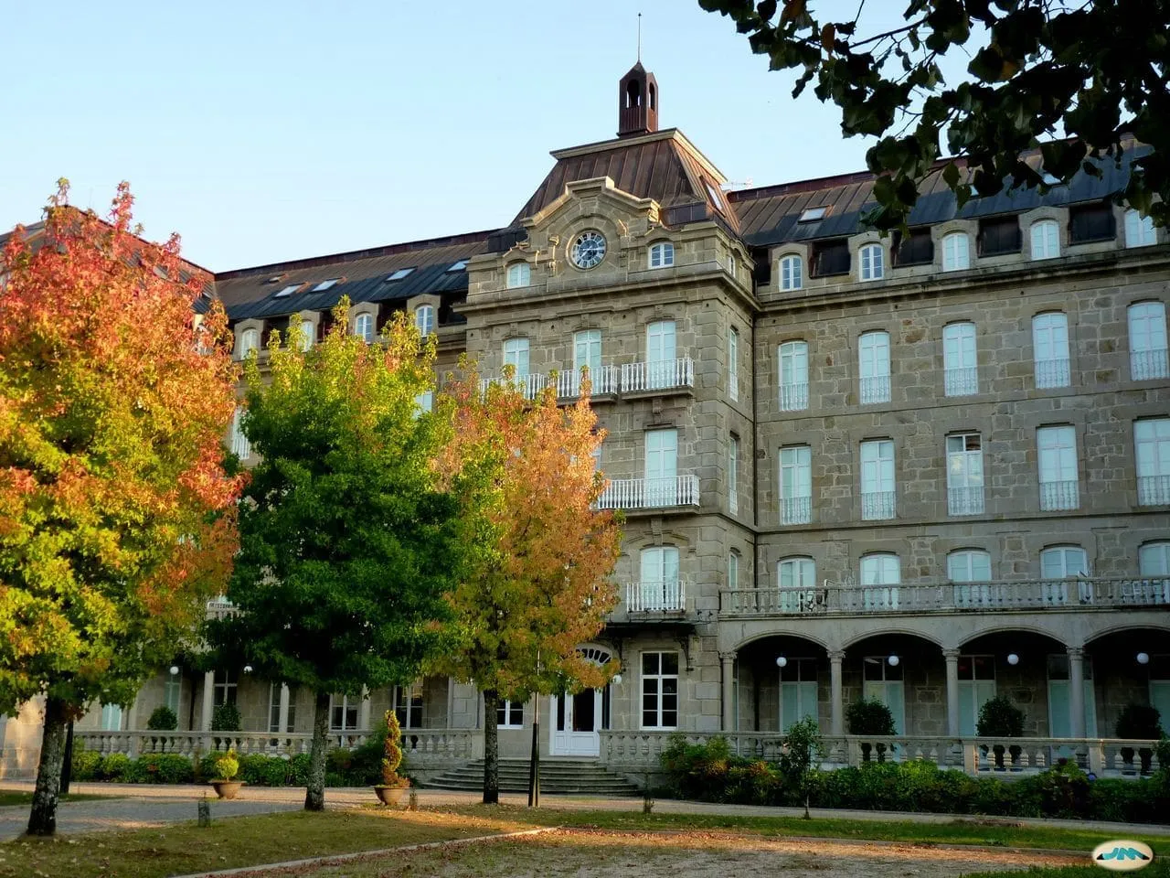 La fachada del Gran Hotel de Mondariz, de piedra, con reloj y tejados de pizarra, tras árboles de otoño