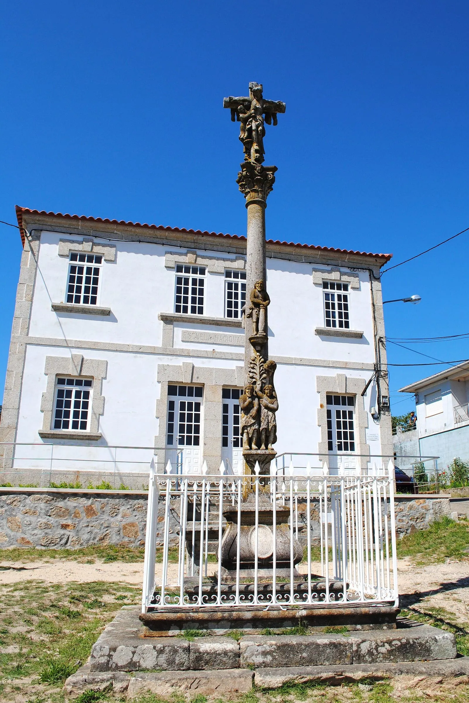 El cruceiro dos Maios, con su fuste esculpido, junto a una casa blanca de ventanas con marco de piedra