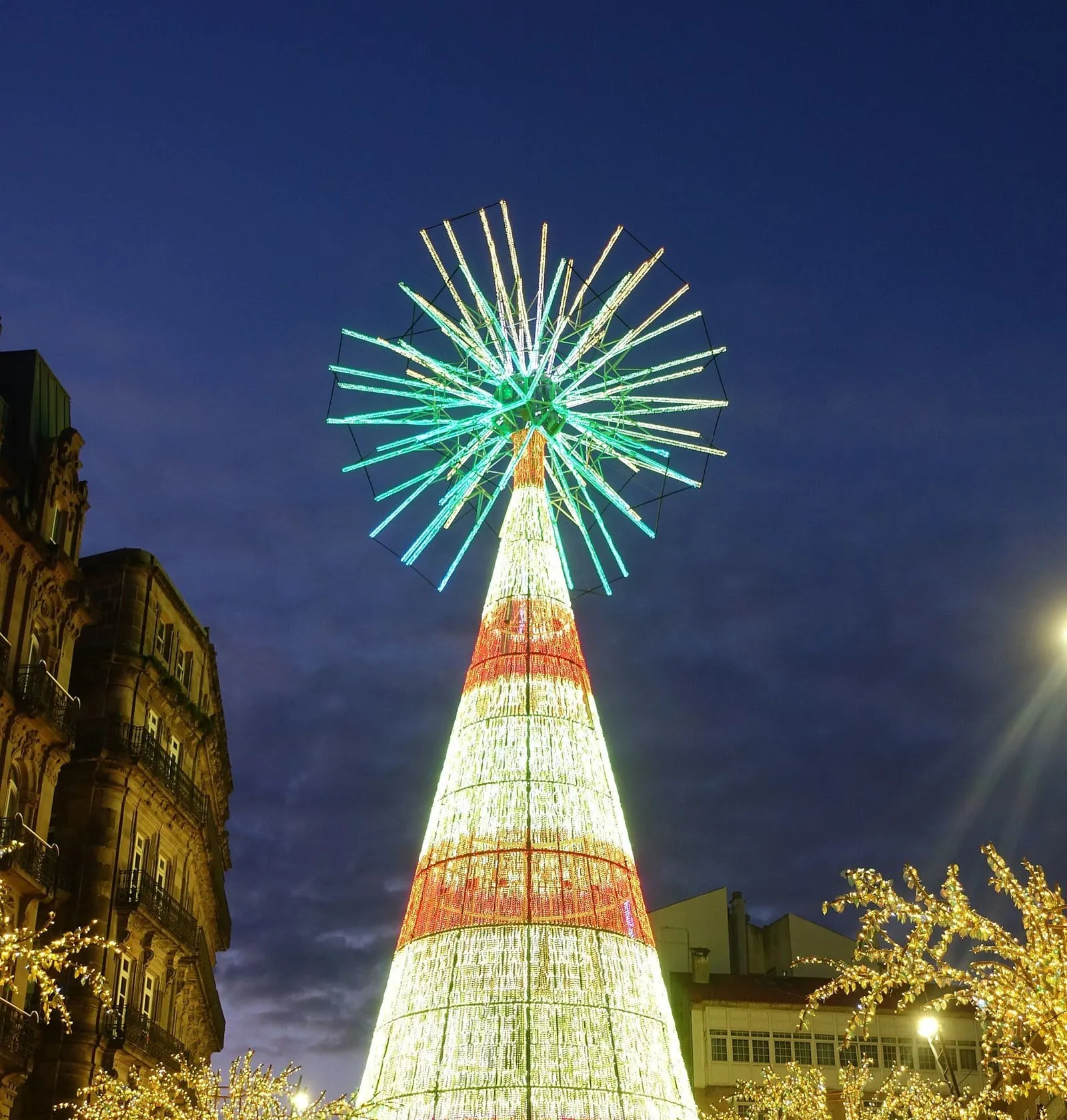 El gran árbol de Navidad iluminado de Vigo, un cono de luces con una estrella en lo alto, entre la multitud al anochecer