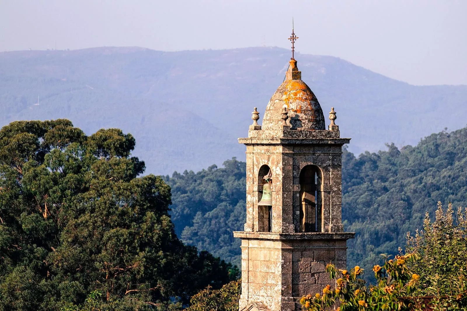 Campanario de piedra de la iglesia de San Tomé de Piñeiro entre árboles, con los montes al fondo