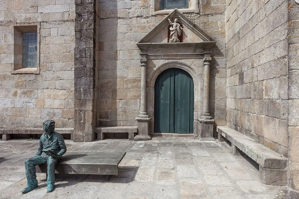 Portada lateral de la iglesia con frontón triangular y una escultura de un niño sentado en un banco de piedra