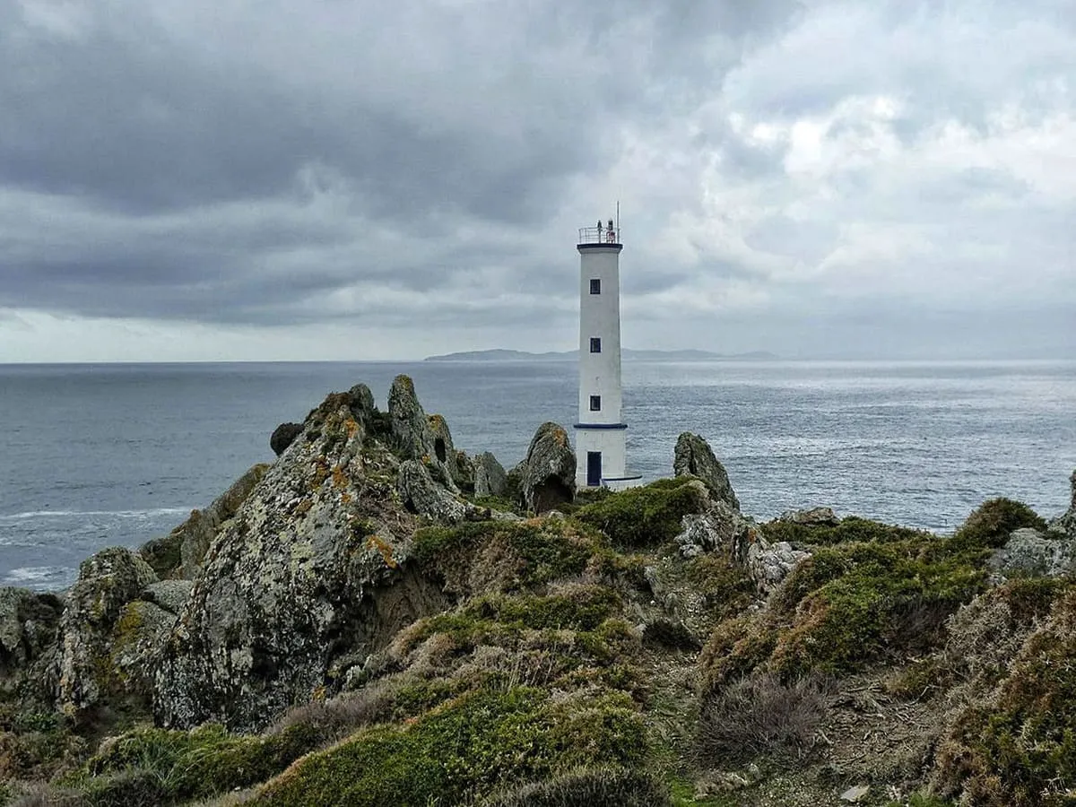 Faro blanco de Cabo Home sobre rocas, con el mar y un cielo nublado