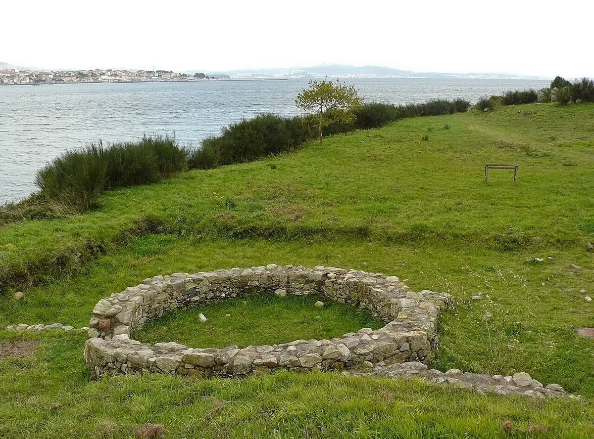 Vivienda circular de piedra en la hierba, con el agua de la ría detrás