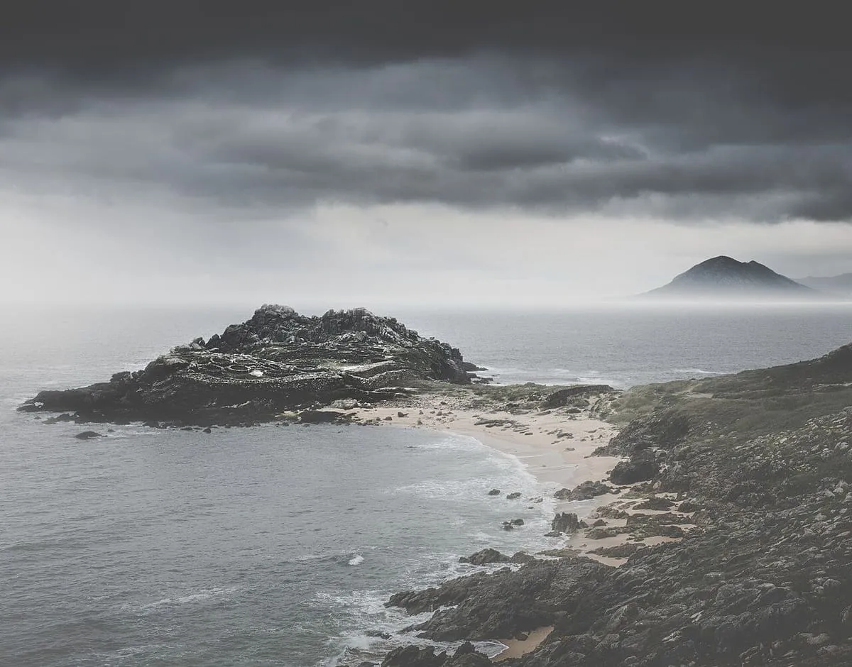 El castro de Baroña en blanco y negro bajo nubes oscuras, con la costa y un monte al fondo
