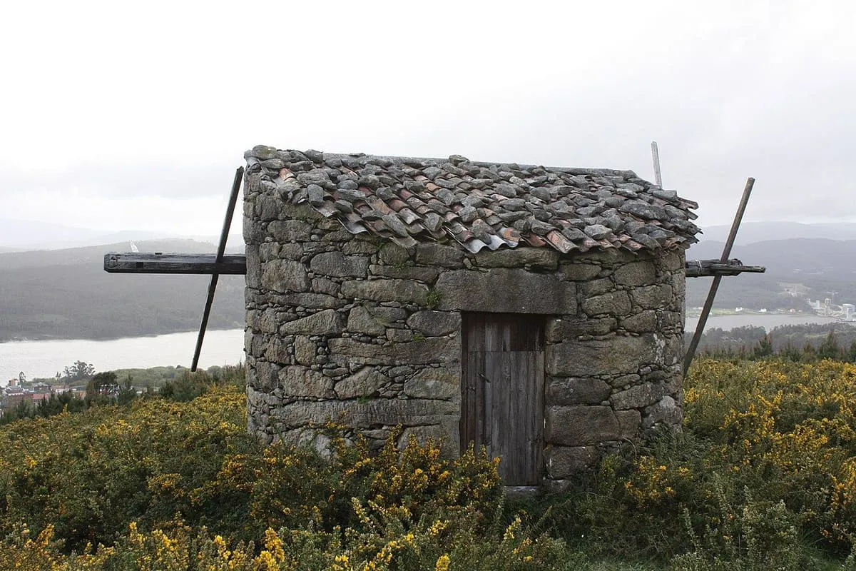 Molino de viento de piedra con el tejado de lajas y las aspas de madera, entre tojos