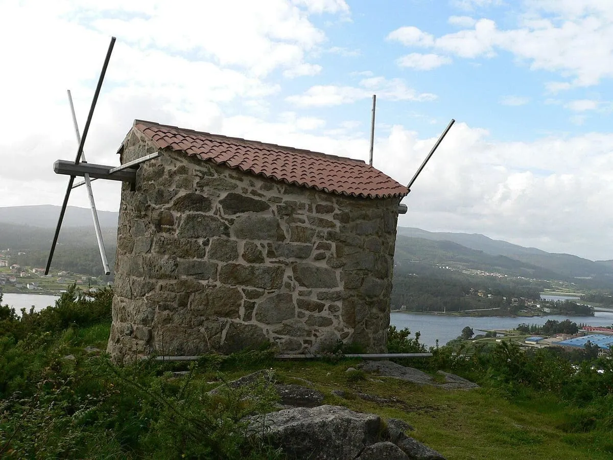 Un molino de viento de piedra con tejado de teja, con la ría del Ulla al fondo