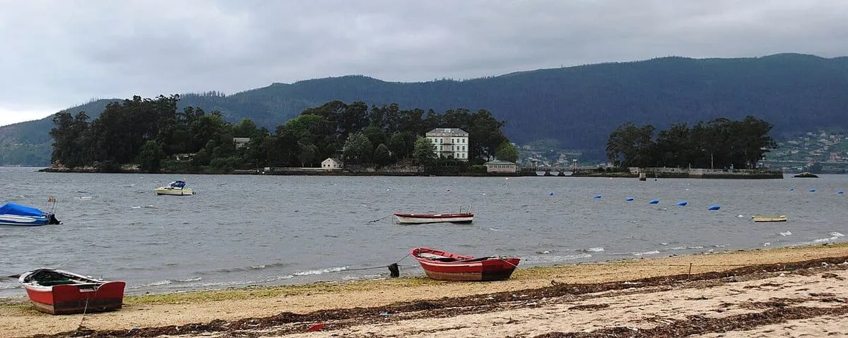 La isla de San Simón, cubierta de arbolado, al fondo de la ensenada, con barcas de remo en la orilla