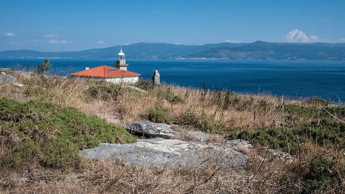 El faro del monte Louro entre rocas y matorral, con la ría de Muros y Noia al fondo