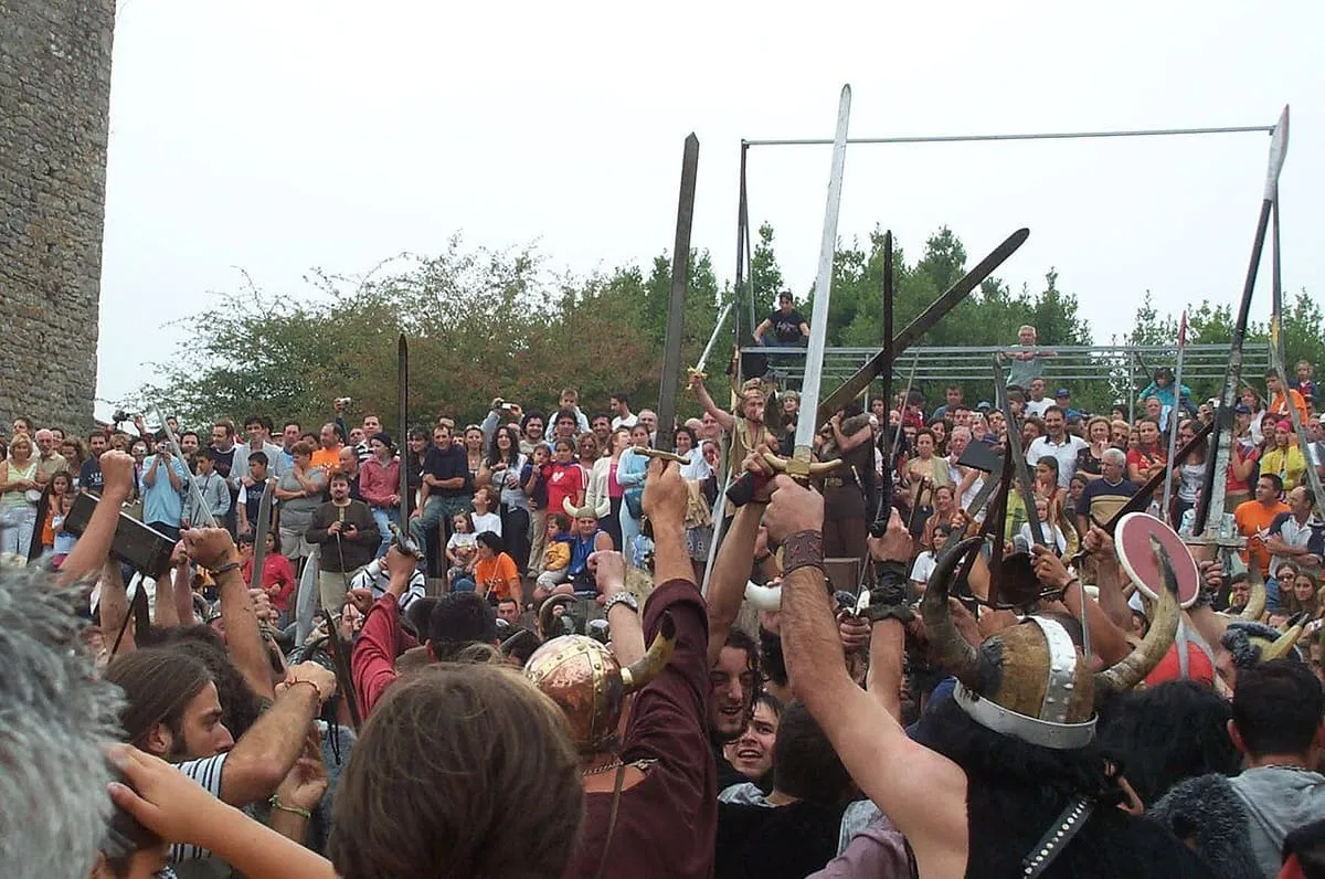 Multitud de participantes de la Romería Vikinga con armas y escudos alzados junto a las torres