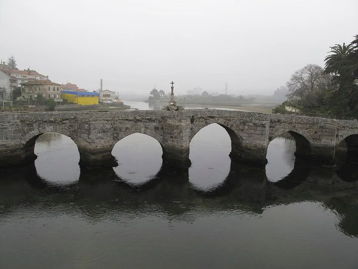 Los arcos del puente medieval de A Ramallosa reflejados en el agua, con el cruceiro al fondo