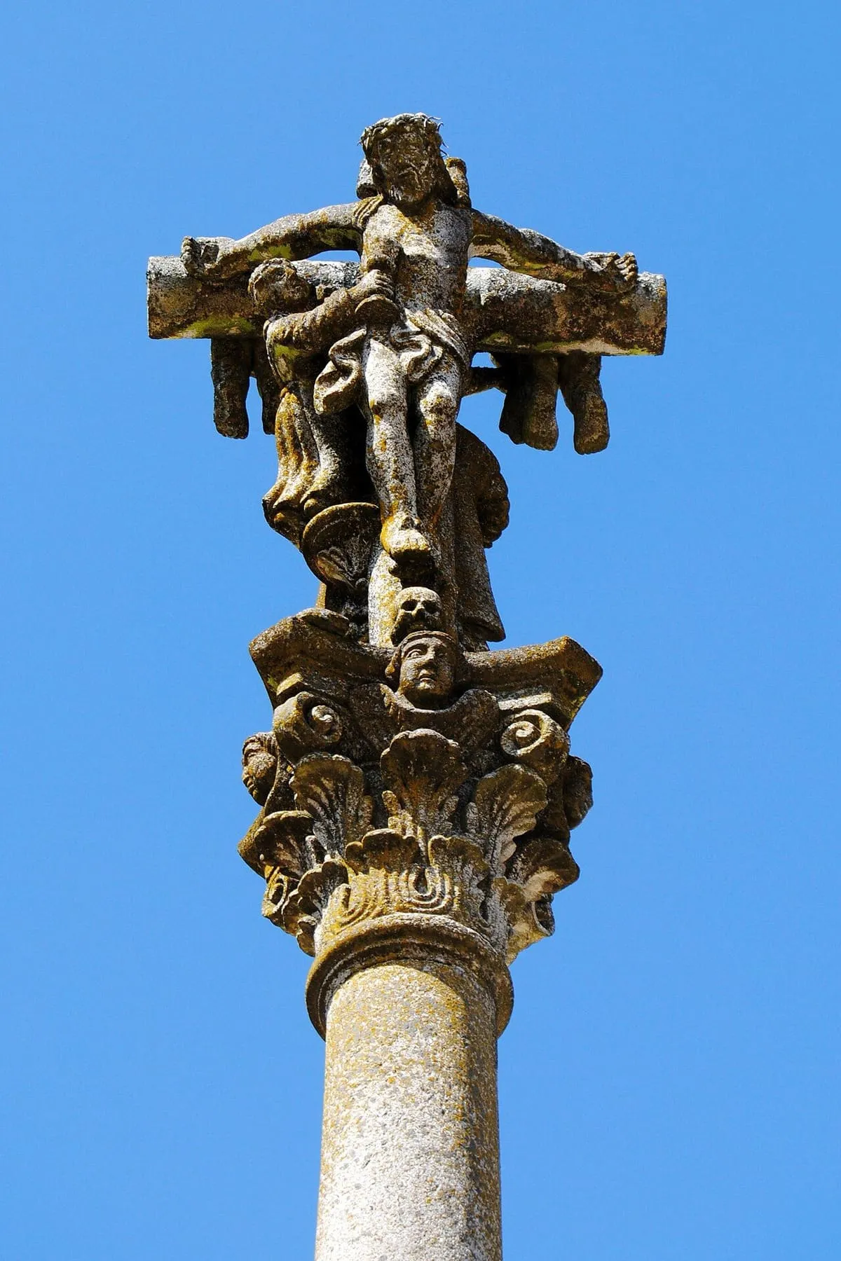 Capitel dórico con hojas de acanto y cabezas de ángel bajo la escultura del cruceiro, sobre cielo azul