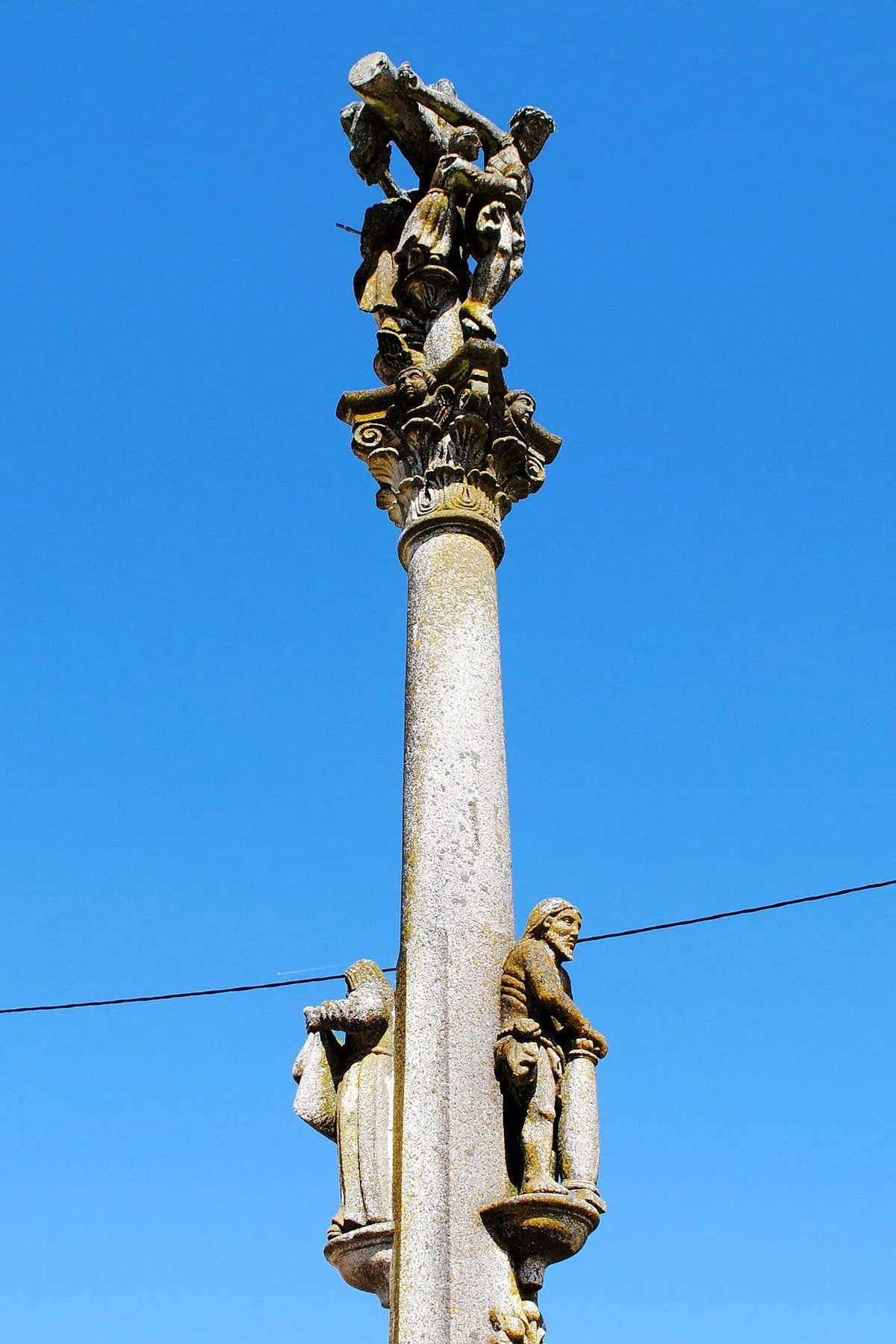 Fuste liso del cruceiro con una figura en la base y la flagelación más arriba, sobre cielo azul