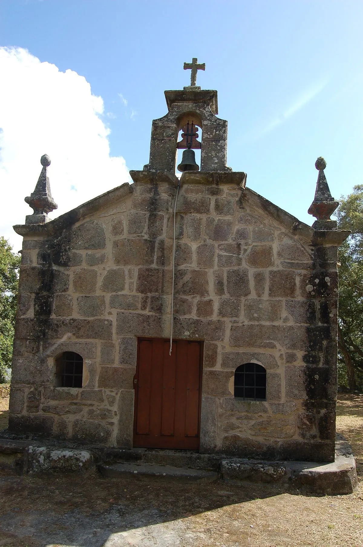 Fachada de piedra de la ermida con una puerta de madera rojiza y dos ventanas pequeñas