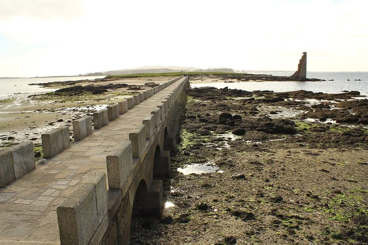 Pasarela de piedra sobre la arena y las rocas de la marea baja, con la torre al fondo