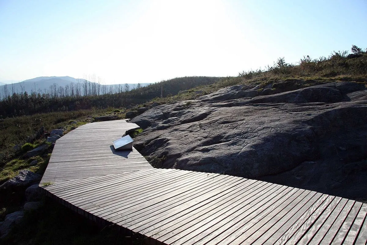 Pasarela de madera entre las rocas de granito del área arqueológica, con montañas al fondo