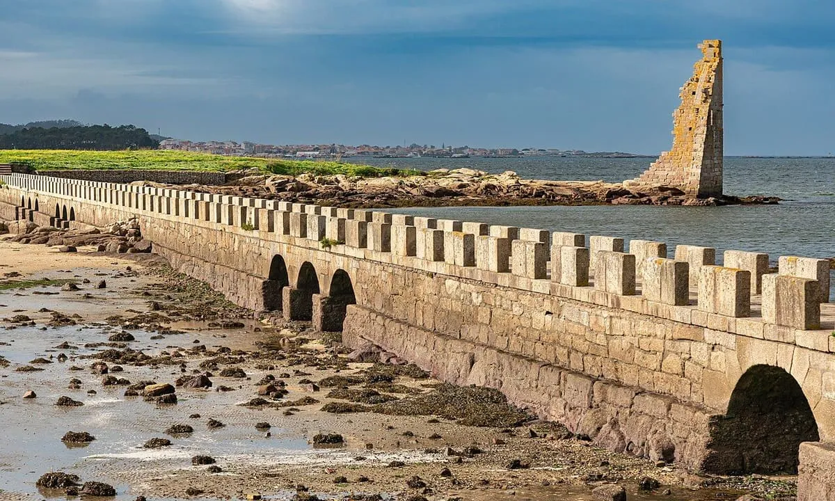 Puente de piedra con arcos y almenillas que cruza la zona de marea hacia la torre de San Sadurniño