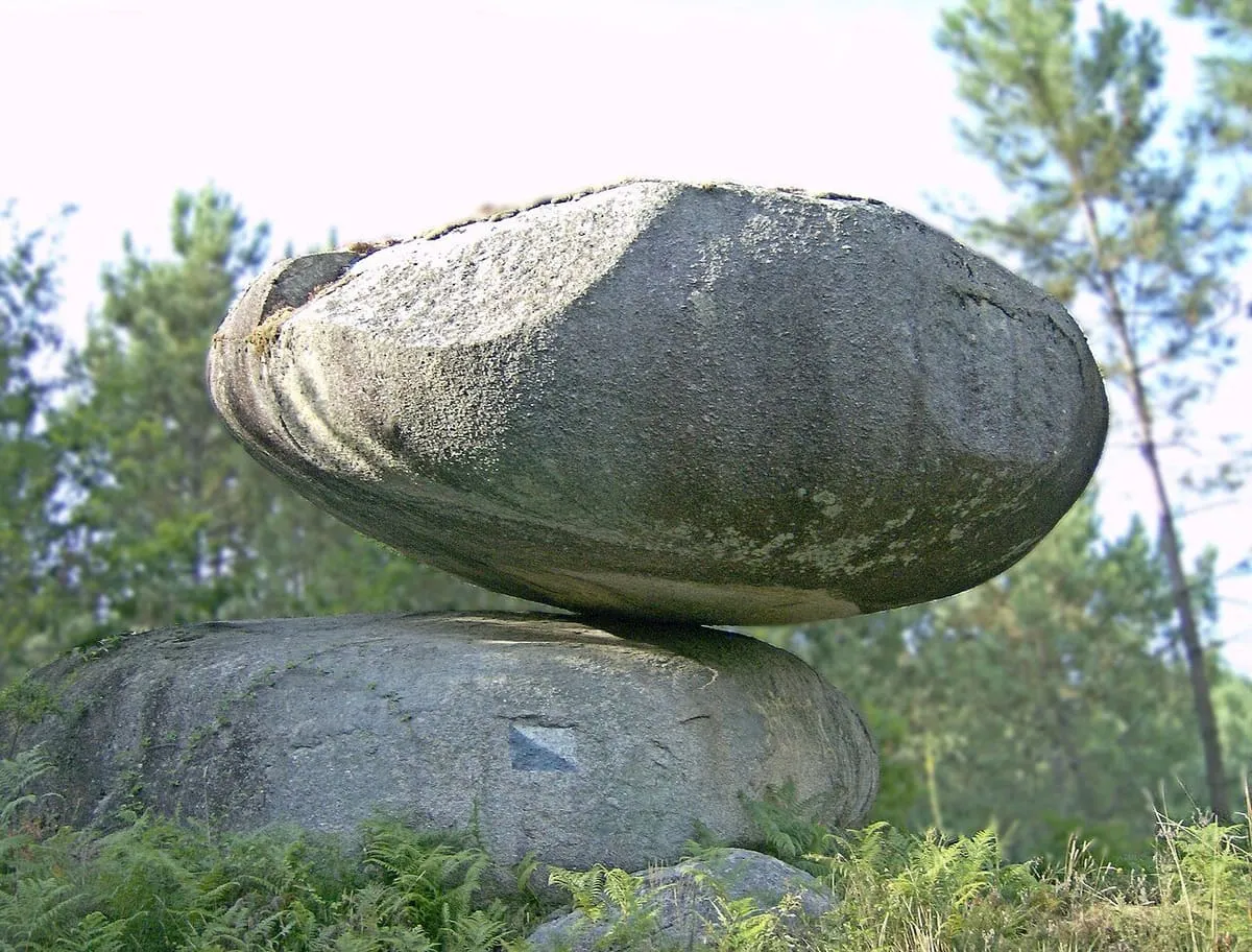 Un enorme penedo de granito liso apoyado sobre otro, entre pinos y matorral