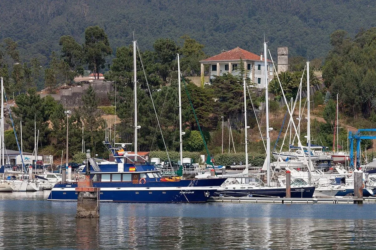 Veleros y barcos de recreo amarrados en el puerto deportivo de Portosín, con un edificio y el monte detrás