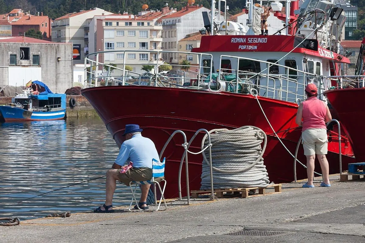 Un barco pesquero rojo amarrado en el muelle de Portosín, con dos personas sentadas junto a las amarras