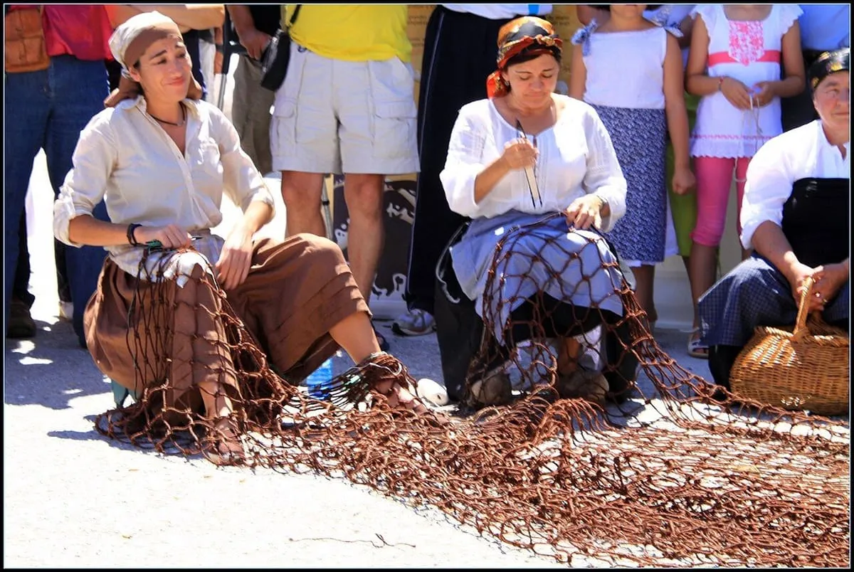 Dos mujeres con ropa tradicional tejen a mano una red de pesca marrón sentadas en la calle