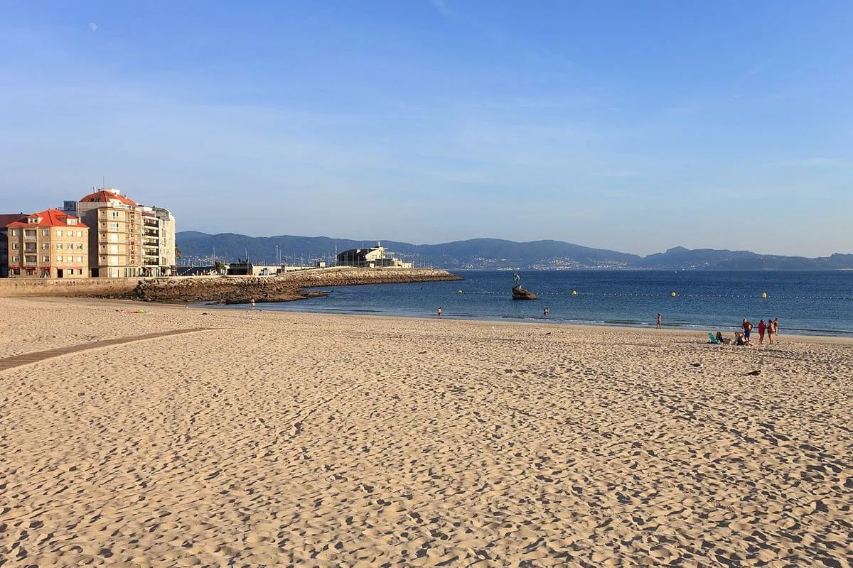 Arena dorada de la playa de Silgar casi vacía por la mañana, con edificios al fondo
