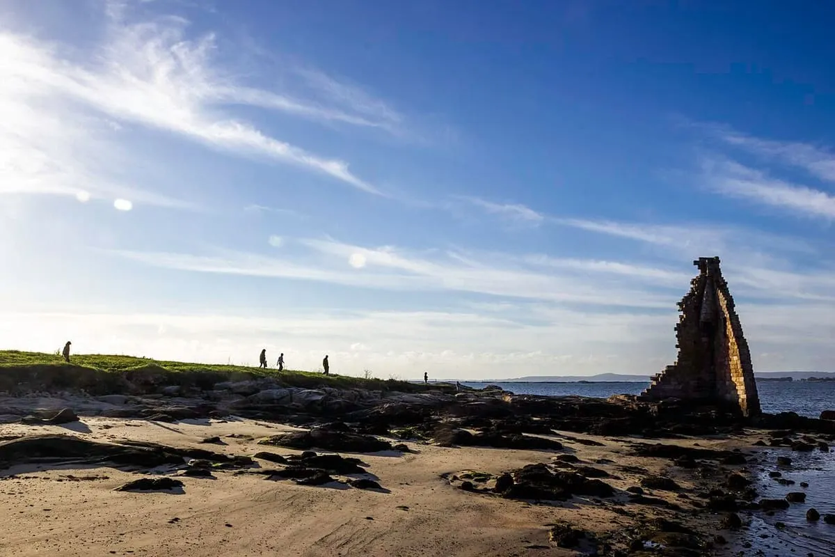 Gente paseando por la arena descubierta con la marea baja, con la torre de San Sadurniño a lo lejos