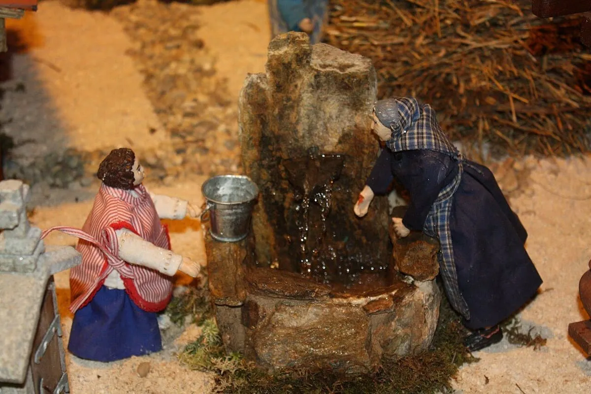 Dos figuras junto a una fuente de piedra con un cubo, en una escena de trabajo del Belén de Valga