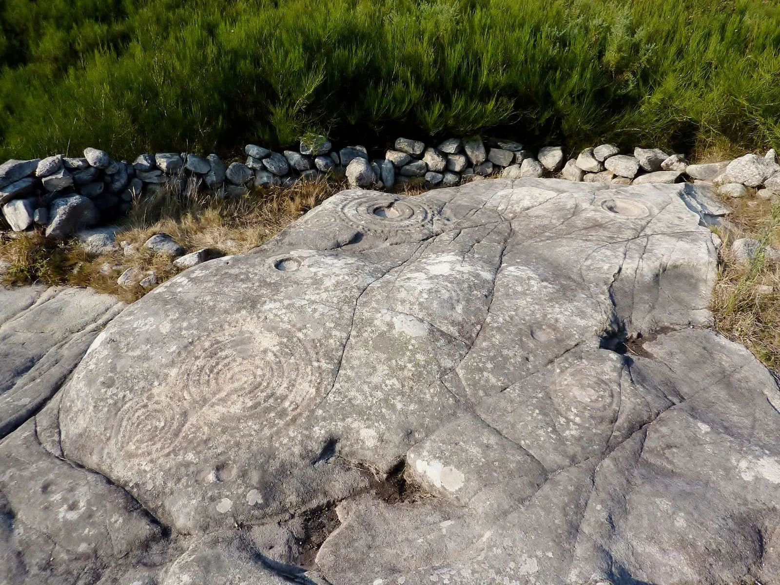 La laxe de granito de Laxe das Rodas con círculos concéntricos grabados, junto a un muro de piedra