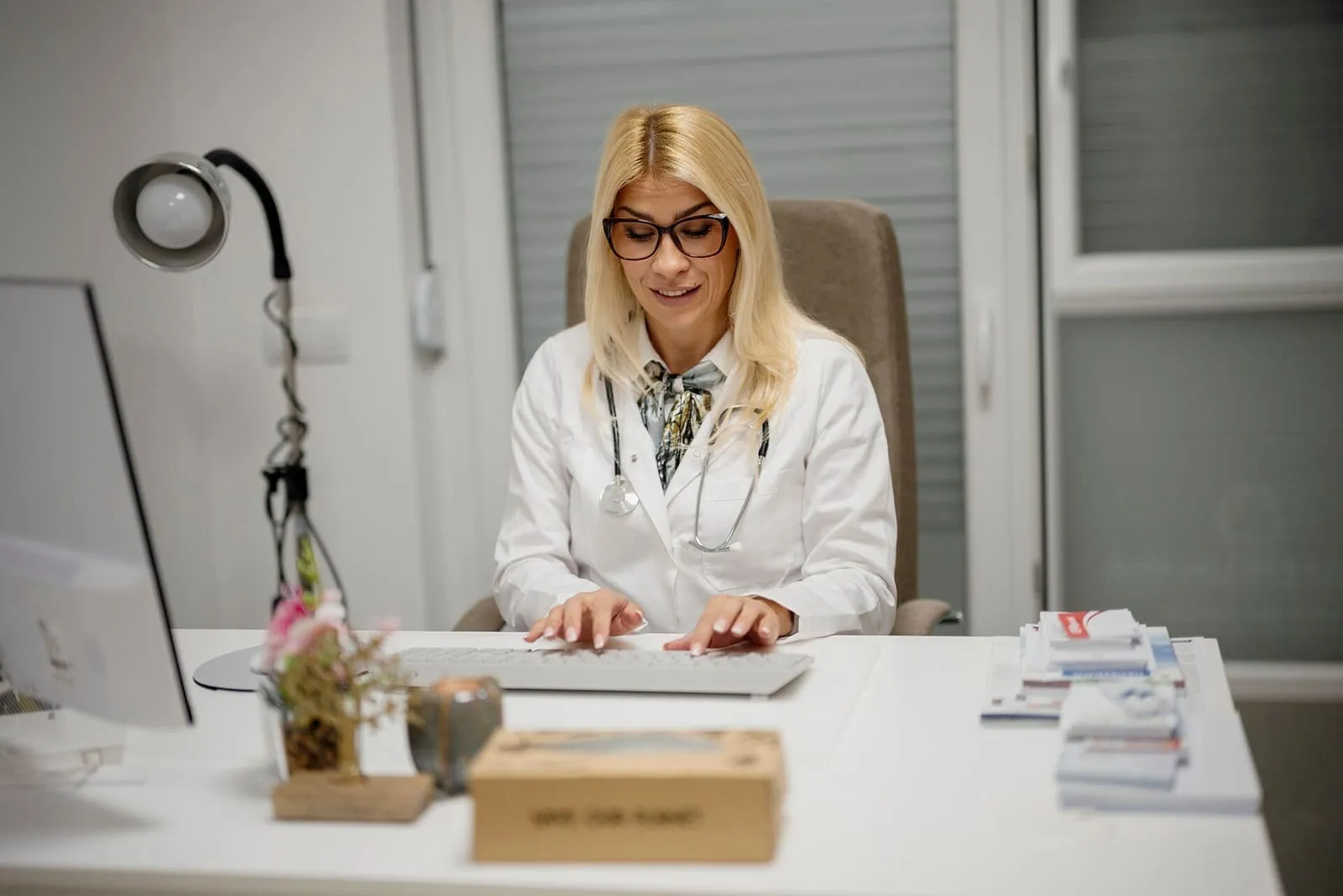Médica con bata blanca y fonendoscopio trabajando con el ordenador en su consulta