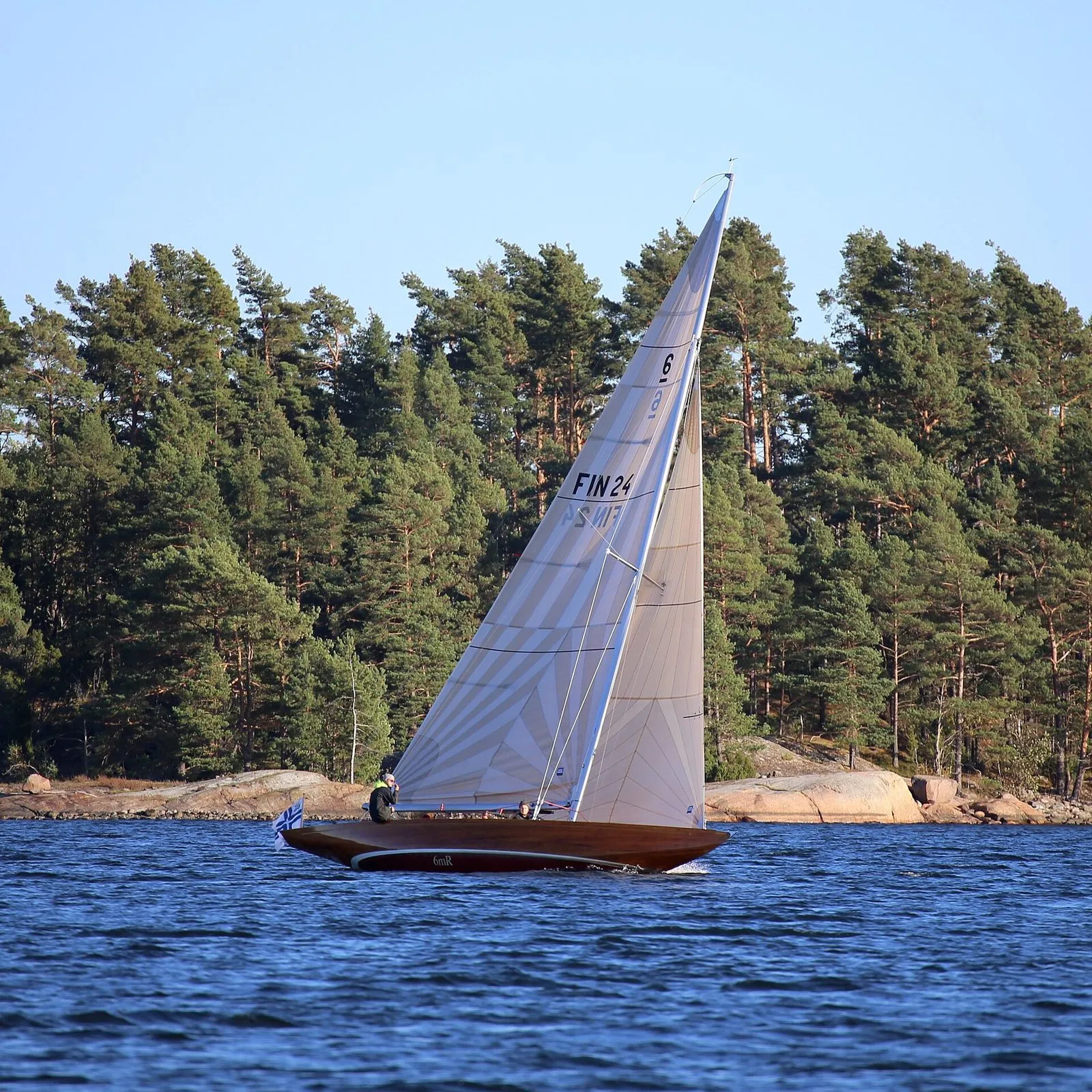 Velero de la clase 6 Metros, con el número 6 en la vela mayor, navegando junto a una costa rocosa; imagen de archivo de la clase