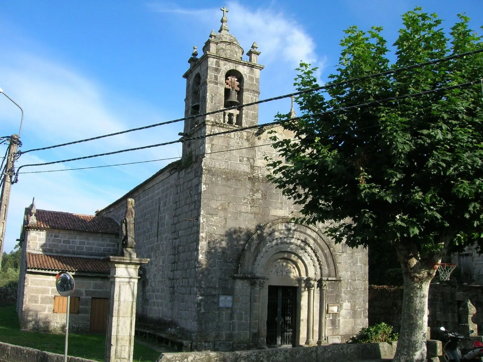La iglesia de Santa Baia de Donas, de piedra, con portada de arquivoltas y campanario de espadaña entre árboles