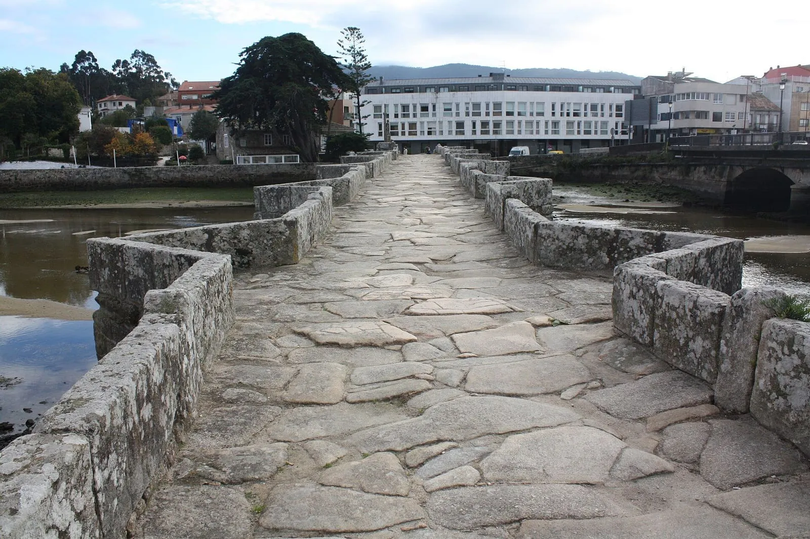 El empedrado del puente medieval de A Ramallosa, entre muretes de piedra, con edificios al fondo