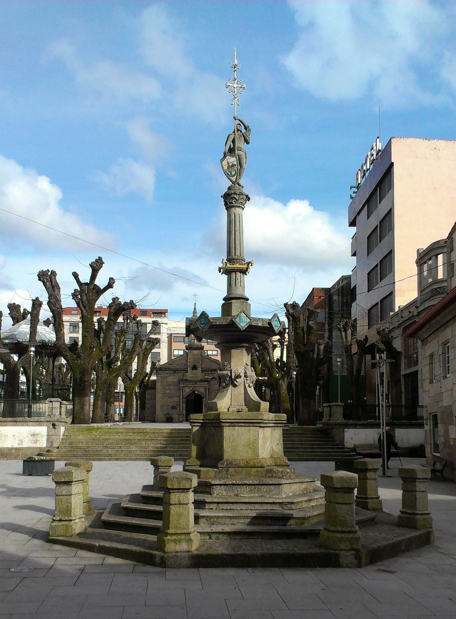 La Fonte do Cristo, con un fuste vertical, en una plaza de O Porriño con edificios y árboles podados