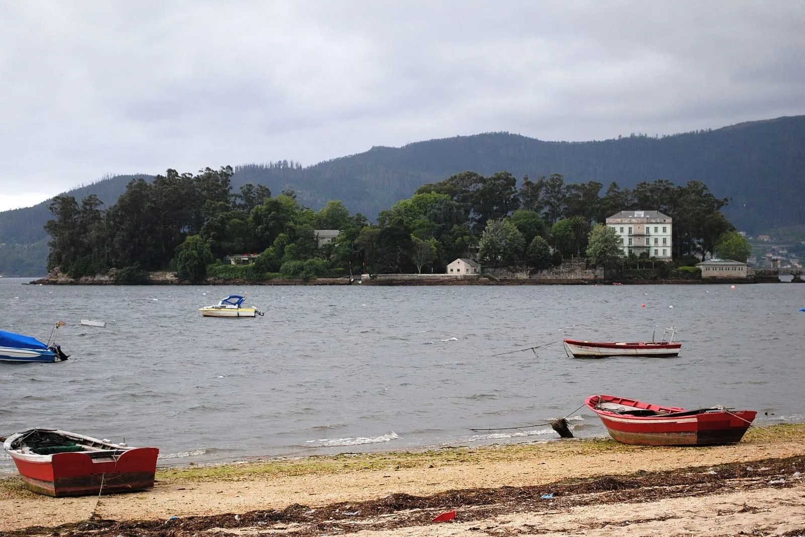 La isla de San Simón, con sus edificios entre árboles, y barcas varadas en la orilla de Cesantes