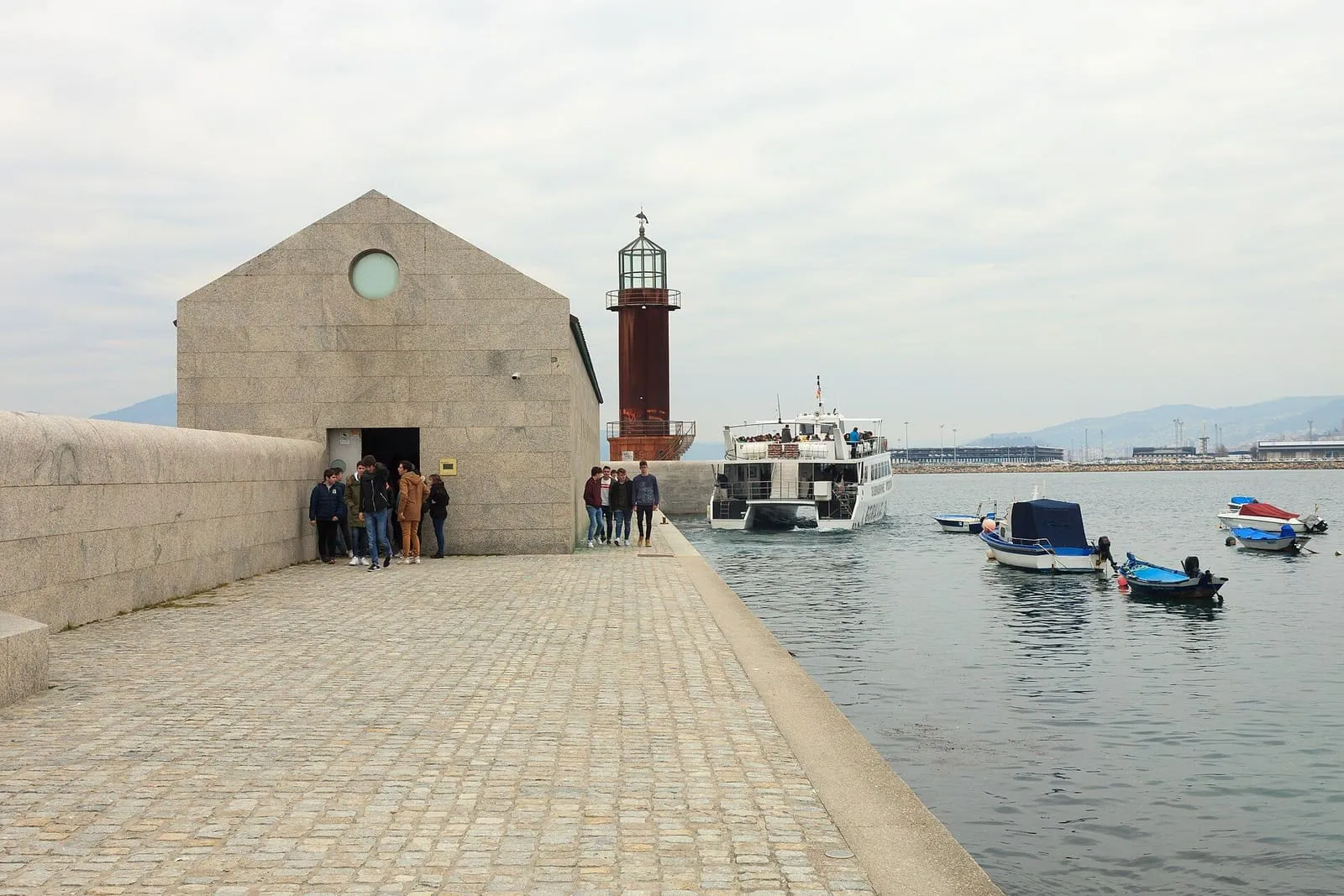 Edificio de piedra del Museo do Mar de Galicia junto al agua, con un faro rojo al fondo
